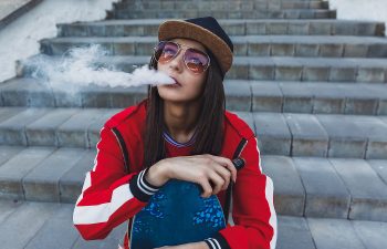 Person sitting on steps, wearing a red jacket and cap, exhaling vapor while holding a skateboard.