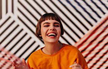 Person in an orange shirt smiling widely, standing in front of a geometric striped background.